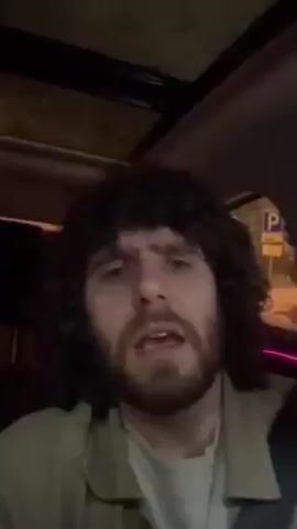 A man with curly brown hair and a beard is in a car. The interior is dark. He's wearing a light-colored jacket over a lighter shirt. He is speaking to the camera. Rain is visible on the sunroof. He looks both at the camera and to the side. He appears to be singing or talking with emotion. The car is moving.