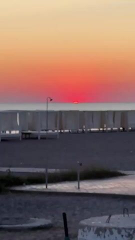 The video shows a sunset over the ocean. The sky is a gradient of orange and pink, with the sun appearing as a bright red disk just above the horizon. In the foreground, there is a beach with lounge chairs and cabanas. There is also a showerhead visible on a pole. The water is calm, and there are a few boats in the distance. The overall scene is peaceful and serene.