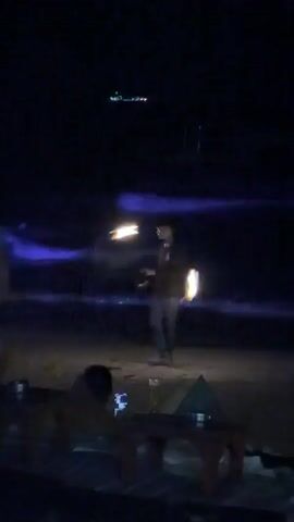 A man juggles burning torches on a beach at night. The man is wearing a cap and dark clothing. The torches are emitting bright flames, illuminating the surrounding area. The background shows a dark sky with some distant lights, possibly from a town or boats. The sea is visible with gentle waves that are illuminated with a purple glow. In the foreground, there are silhouettes of people sitting at a table, with drinks on it. The overall scene is dark and atmospheric, with the fire performance being the main focus.