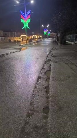 The video shows a dark street at night after a rainfall, reflected by the wet pavement. The street is lined with streetlights, some decorated with brightly colored, stylized floral designs and heart-shaped lights. The camera moves forward, showing the crosswalk with yellow and white stripes. The video then points up at a very bright streetlight before going black.