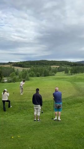 The video shows a golfer in mid-swing, driving a golf ball on a green golf course. Three other individuals are present, observing the golfer. One person is standing with their back to the camera holding a golf club. Another person is standing with their back to the camera and another individual is off to the left, taking a photo with their phone. The surrounding environment includes rolling green hills and trees, under an overcast sky.
