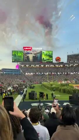The video shows a large crowd of people gathered at Levi's Stadium. A large screen displays images, and fireworks are exploding in the sky. Many people are holding up their phones, presumably to record the event. The crowd appears dense, and individuals are dressed in various clothing. The field is visible, and there are some structures or equipment on it. The sky is partly cloudy.