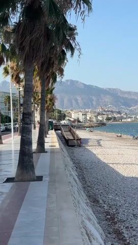 The video shows a view of a beach with palm trees in the foreground and a town in the background. The beach is made of small white pebbles. There are a few people sitting on the beach under umbrellas. The sea is calm and blue. In the distance, there are mountains. The sky is clear and blue. The video pans from left to right, showing more of the beach and the town. There is a walkway with a ramp leading down to the beach. There are also some cars parked on the left side of the walkway. The overall impression is a peaceful and relaxing beach scene.