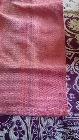 The video shows a close-up of a pink woven fabric that appears to be a rug or a mat. The fabric has a textured surface with visible threads. The right side of the fabric is slightly curled or folded over. Beneath the pink fabric, a patterned cloth with purple and white designs is visible. The lighting in the video is somewhat diffused. There are no people or significant actions taking place in the video, it is a static shot of the fabric.