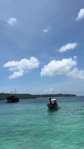 Two boats are visible on turquoise water under a blue sky with scattered white clouds. The boat in the foreground is a long, narrow vessel with a canopy, a red and green decorative element at the front, and a motor. The boat in the background is a larger, dark-colored vessel resembling a pirate ship, with masts and rigging. A distant green, tree-covered island with buildings is visible on the horizon. The water appears clear and calm, with a rope visible in the lower right corner of the frame. The lighting suggests it is daytime.