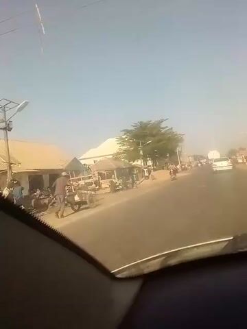 The view from a car driving down a two-lane street is shown in the video. The sky is clear and blue. On the left side of the road are small buildings with corrugated metal roofs and people standing outside. A pile of dirt sits near a light-colored building. Trees provide some shade. A motorcyclist is approaching from the opposite direction, and a white car is driving in the distance in the same direction as the camera. The dashboard of the car is visible at the bottom of the frame. The scene appears to be set in a bustling commercial area with a mix of pedestrians and vehicular traffic.