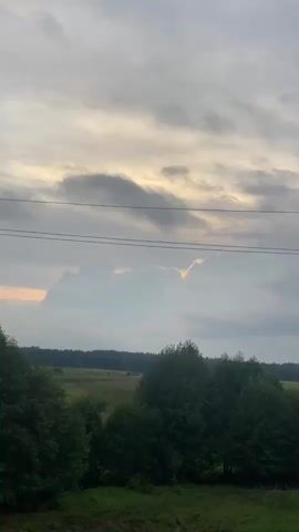 The video captures an outdoor scene with a cloudy sky. In the foreground, there is a large concrete utility pole with wires stretching across the frame. The pole has insulators on top. Trees with dense green foliage are visible on either side of the pole, and a field with some distant trees can be seen in the background. The clouds appear dramatic, with shafts of sunlight breaking through in some areas, creating bright highlights. There are no people or animals visible in the video. The overall impression is a rural landscape under a dynamic sky.