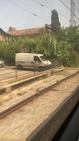 The video shows a view from a moving train. The camera is positioned to look out the right side of the train. In the foreground, there are train tracks and ballast. Beyond the tracks, there is a landscape with trees, bushes, and some buildings in the distance. There are also overhead power lines and utility poles visible. The lighting suggests it is daytime, likely sunny. The movement of the train causes a slight blur to the scenery.
