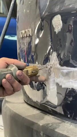 The video shows a person's hands using a dent puller tool on a damaged section of a dark-colored Toyota vehicle. The tool is being applied to areas where the paint has been removed and metal is exposed. The damaged area appears to be on the lower rear quarter panel, near the bumper. The environment seems to be a repair shop, with another vehicle visible in the background.
