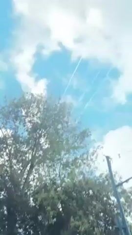 The camera is pointed upwards towards a partly cloudy blue sky. There are three white contrails streaking diagonally across the sky from upper left to lower right. In the foreground is a green tree, its leaves obscuring some of the sky. There are also power lines visible on the right side of the frame.
