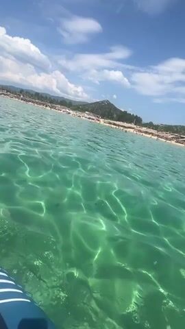 The video begins with a close-up of a turquoise ocean with sunlight patterns on the surface. A blue paddle is visible in the lower right corner. The camera then pans across the water, revealing a crowded beach with many people and umbrellas in the background, bordered by a line of trees and hills under a bright blue sky with scattered white clouds. The perspective suggests the camera is on a paddleboard or similar watercraft, as a person's bare foot is visible in the lower left corner of the frame from 00:02 to 00:04. The water is clear, and the seabed is faintly visible beneath the shimmering surface.