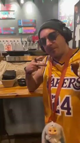 The video shows a man indoors, wearing a yellow Lakers jersey with the number 24 on it, a gray beanie, sunglasses, and headphones. He holds a sandwich in his right hand and a small stuffed bunny on an orange lanyard around his neck. He poses for the camera, occasionally pointing. There is a bar behind him with drink taps and decorations. The background music is upbeat.