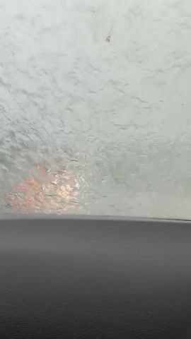 The video shows a view from inside a car during heavy rain. Water is streaming down the windshield, obscuring the view of the road ahead. The wipers are in motion, attempting to clear the glass. Trees and other foliage can be seen blurred on either side of the road. The weather appears overcast and the lighting is dim. There are other vehicles visible in the distance, their headlights appearing as hazy glows through the rain. The overall impression is of driving in challenging weather conditions.