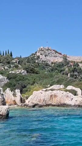 This video captures a scenic coastal panorama from the perspective of a boat on a bright, sunny day in Demre, Turkey.

The scene opens with a magnificent stone fortress or castle positioned atop a rugged, green-covered hill. A Turkish flag is visible flying from within the castle's ancient walls. Below the fortress, a charming town unfolds on the hillside, characterized by numerous houses with distinctive red-tiled roofs nestled among lush vegetation.

As the camera pans right, more of the picturesque town and its structures come into view along the verdant coastline. The foreground is dominated by the crystal-clear, vibrant turquoise and blue waters of the sea. The panning motion reveals a small bay or harbor where several boats, including traditional sailboats with visible masts, are moored. One boat clearly displays a Turkish flag.

Continuing its sweep, the camera shifts focus from the town and harbor towards the expansive open sea. The metal railing of the filming boat's bow appears in the lower right of the frame, indicating the viewer's position on the water. In the distance, a serene line of green, hilly coastline stretches along the horizon under a perfectly clear blue sky. The water is gently rippling, creating a tranquil atmosphere. No specific text is visible apart from the flags. The overall impression is one of a beautiful and historic Mediterranean setting.