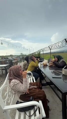 Four women are seated at an outdoor table, eating. The woman on the far left is wearing a brown hijab and a striped shirt. The woman next to her is wearing a yellow top and a dark hijab. The woman in the center is wearing a dark hijab and a dark top. The woman on the far right is wearing a light pink hijab and a maroon top. The table is black and has a shiny surface. There are bags of food and drinks on the table. The women appear to be in a casual setting, possibly a cafe or restaurant, with a fence and a field in the background. The sky is cloudy.
