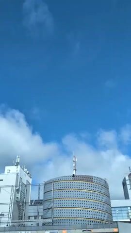 A short, vertical video shows a large, cylindrical building with a textured, metallic facade that has a pattern of small, dark dots. The building is topped with antennas and situated under a bright blue sky with scattered white clouds. To the left of the cylindrical building, a portion of a white, angular structure is visible, also with antennas. To the right, another building, appearing to be made of glass or metal, is partially in view. The video appears to be filmed from a low angle, looking upwards.
