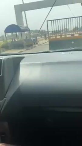 The video begins inside a car, likely a Toyota, as it approaches an entrance gate. A person opens the gate. The camera shifts slightly, revealing the driver who is holding and looking at a red phone. The interior of the car is visible, including the dashboard, steering wheel, and radio.  The environment outside the car shows a building and a street with other vehicles.