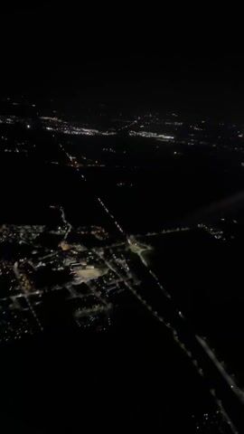 The video captures an aerial view of a landscape at night, likely filmed from an aircraft. Below, a network of artificial lights illuminates the dark ground, revealing a sprawling town or city. Various structures, including large industrial or commercial buildings with well-lit parking areas and rooftops, are visible. Roads crisscross the terrain, marked by streams of vehicle lights.

A prominent red light beam projects upwards into the night sky from a point on the ground. Adjacent to this, a very bright white light source is also visible, casting a strong glow and appearing almost like a powerful spotlight. These bright lights are concentrated in the upper right portion of the frame.

The scene suggests a clear night with good visibility of the ground and atmospheric light effects. No specific text or identifiable landmarks are visible within the video itself. The context provided indicates the location is Carpiano, Italy. The video focuses on the interplay of artificial light against the nocturnal backdrop.