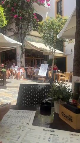 The video shows an outdoor seating area of a cafe. In the foreground, on a table, is a menu, a black ashtray, a small potted plant, a wooden box containing condiments (bottles of what appear to be olive oil and vinegar, salt and pepper shakers, and napkins), and a branded bag. The table is made of dark material, possibly wicker. In the mid-ground, several tables with chairs are occupied by patrons. Some people are seated and talking, while others appear to be looking at their phones. A whiteboard on an easel is visible. A tree trunk is in the center of the frame, with a large white umbrella overhead. The background shows a white building with yellow trim and doorways, some with arched entrances. Floral elements are visible on the buildings and surrounding the area. The scene appears to be illuminated by natural sunlight.