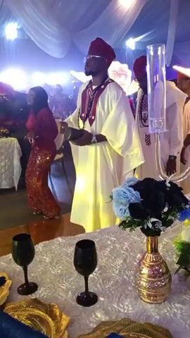 A formal event is taking place, possibly a wedding or reception, with a man in a white robe and a red hat standing center frame. He appears to be holding something in his hands, possibly a book or a tablet, and is looking off to the side. Another man, also in a similar traditional outfit, stands to his right, facing him and gesturing with his hands. In the background, a woman in a red sequined dress is visible, dancing or moving. The environment is decorated with white drapery, chandeliers, and floral arrangements. The lighting is dim and appears to be stage lighting, with colored lights illuminating the background. There are tables set with white tablecloths, gold plates, and black wine glasses. The overall atmosphere suggests a celebratory occasion.