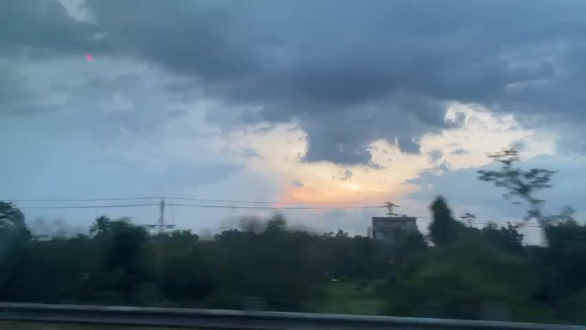 The video shows a blurry, time-lapse view of a cloudy sky at dusk, possibly during a storm, as seen from a moving vehicle. The sun is setting, with orange and pink hues visible through gaps in the dark, heavy clouds. The landscape below consists of trees, power lines, and some distant lights. The motion blur indicates the camera is in transit.