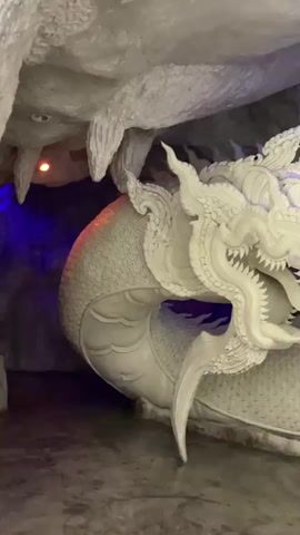 A white dragon statue with a red eye is visible in a cave-like setting. The statue is detailed, with intricate carvings along its body and head. The cave walls appear rough and textured. There are colored lights, blue and green, illuminating parts of the scene. The camera pans and zooms, focusing on different parts of the statue.