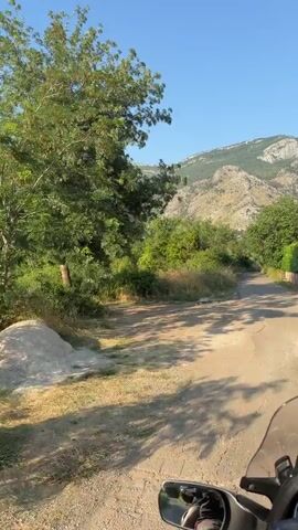 The video shows a first-person perspective from a motorcycle traveling on a rural road in Buljarica, Montenegro. The road is paved but shows signs of wear, with some patches of dirt and gravel. Lush green foliage and trees line both sides of the road, with a dense cluster of vegetation on the left and a wooden fence visible on the right. In the background, a majestic mountain range with rocky, arid slopes rises against a clear blue sky. The sun is shining brightly, casting long shadows from the trees across the road, indicating that it is likely morning or late afternoon. The overall atmosphere is one of peaceful exploration and scenic beauty. The camera is mounted on the motorcycle, capturing the handlebars, a rearview mirror reflecting a person's arm and shoulder, and a portion of the dashboard. There is no visible text in the video.