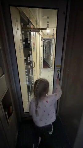 A young child with braided blonde hair presses a button to open an automatic glass door inside a train. The child is wearing a long-sleeved shirt with a pattern and dark-colored pants with white polka dots, as well as white sneakers. The door has a frosted pattern on the glass and a sign in Russian above it.  The hallway beyond the door is visible, with beige walls and a shiny floor. The train car appears to be dimly lit.