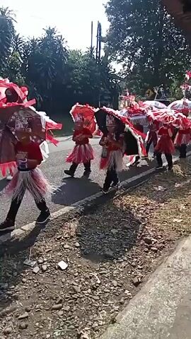 A group of children, dressed in red and white outfits and carrying large, decorative umbrellas, walk in a procession along a paved road. Some of the umbrellas are red and white, while others are blue. The children are walking on the right side of the road, with trees and foliage visible on the left. Several motorcycles are also present, with one person riding a motorcycle in the procession. The scene appears to be a parade or celebration, possibly in a tropical or subtropical region given the lush vegetation. The ground beside the road is covered in gravel and debris. The overall mood seems festive.