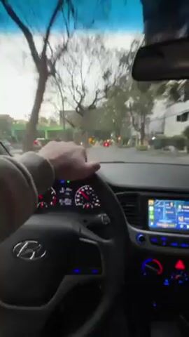 The video shows a first-person perspective from inside a car, presumably driven by the person recording. The car, identified by the Hyundai logo on the steering wheel, is moving through a city street. The person's hand is visible on the steering wheel. The dashboard shows a digital display with vehicle information, and a touchscreen displays a navigation map. Streetlights and traffic lights are seen through the windshield, illuminating the road ahead. Trees line the street on both sides, suggesting a residential or suburban area.