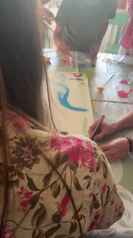 Several people are gathered around a table covered with a tablecloth with a light green background and a circular pattern in white. They are working together to create a large painting on a white sheet of paper. The painting includes elements like a blue river, abstract shapes in red, and brown mountains.  The word "CIVILISATIA" is written in green on the right side of the paper, and someone is adding the letter "P" in red next to it. There are various containers with paint, brushes, and markers scattered across the table. One person is wearing a floral patterned shirt with pink and brown colors, while another wears a striped gray and white shirt.