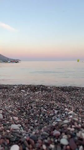 The video captures a low-angle, close-up view of a pebble beach with the sea and a pastel-colored sky in the background. The pebbles in the foreground are varied in size and color, with shades of gray, brown, and white. Small waves gently lap the shore, creating a shimmering effect on the water's surface. In the distance, a calm sea stretches to the horizon, meeting a sky painted with soft hues of pink, purple, and blue, indicative of either dawn or dusk. A faint line of land or hills is visible on the far left, and a yellow buoy floats in the water on the right. There are no people or animals visible in the frame. The overall atmosphere is peaceful and serene.