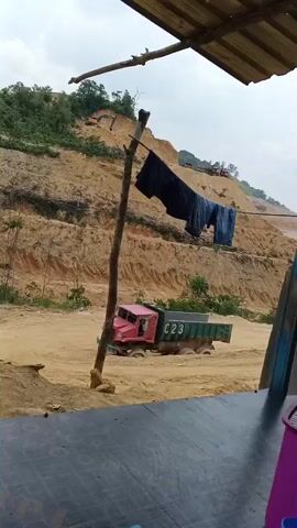 A red dump truck with a green bed is stuck in the mud at a construction site. The truck has the number C23 on its side. In the background, there is a large hill with terraces carved into it, and an excavator is visible on the hill. Clothes are hanging on a clothesline near a wooden pole in the foreground. The ground is covered in dirt and sand. The sky is overcast.