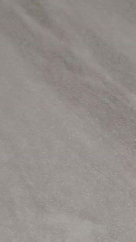 The video begins with a close-up of a gray patterned surface. The camera then pans upwards to reveal the seam between the gray surface and a beige tiled surface. The camera continues to pan upwards, showing a grid of white supports below the tiles. The video concludes with a person's hand in a patterned sleeve gently pushing the gray surface upwards. The environment appears to be an interior space with unfinished construction or renovation.