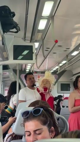 The video shows the interior of a train. The camera is positioned in the aisle, looking forward. In the foreground, two women are seated, one with sunglasses on her head and the other in a red dress. In the middle ground, a man in a white t-shirt stands next to a large blonde puppet dressed in a red robe. The puppet's mouth is open, and it appears to be moving. Other passengers are visible in the background, some seated and some standing. The train appears to be in motion, with a view of greenery and the sea through the windows on the right side. The lighting inside the train is provided by fluorescent tubes overhead.