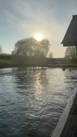 The video shows a long pool of water outdoors, with several people swimming in it. The sun is shining brightly, creating reflections on the water. A tree is visible in the background. A building with a dark roof and windows is partially visible on the right side of the frame, next to the pool. The camera slowly moves from left to right, providing a view of the pool and the surrounding area.