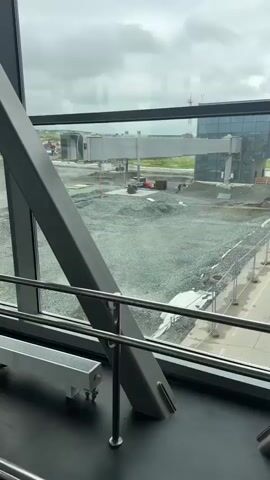 The video pans across a large window showing an airport gate under construction. The window is framed by dark grey metal beams. Outside, a jet bridge extends from the terminal building to a construction area. Piles of gravel and construction equipment, including an excavator and trucks, are visible. The sky is overcast.