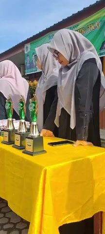 The video shows a close-up view of a table covered with a yellow tablecloth. On the table are several trophies lined up, all identical in design with green and gold accents. Behind the table, three women wearing hijabs and black garments are standing. The woman in the foreground, on the right, is looking down at the table and appears to be interacting with something on it, possibly a phone. Another woman, in the middle, is also looking downwards. A third woman, on the far left, is partially visible and facing away from the camera. In the background, a banner is visible with text that seems to indicate an event or competition. The setting appears to be outdoors, with natural light suggesting daytime.