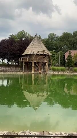The video displays a wooden watermill structure situated beside a green body of water. The watermill features a large wooden wheel. The water reflects the structure and the surrounding trees. The sky appears overcast. In the foreground, railroad tracks are visible.