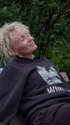 A woman with curly blonde hair, wearing a black sweatshirt with a camouflage pattern and the word "WINNER" on it, is sitting outside on what appears to be a bench. She closes her eyes, tilts her head back, opens her eyes, and then begins to talk. A bottle of wine with a red stopper is visible behind her. The background consists of green trees and foliage.