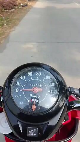 A first-person perspective video shows a motorcycle dashboard with a speedometer indicating a speed of 20 km/h. The road ahead is visible, lined with trees and some vegetation. The motorcycle appears to be in motion on a paved road.