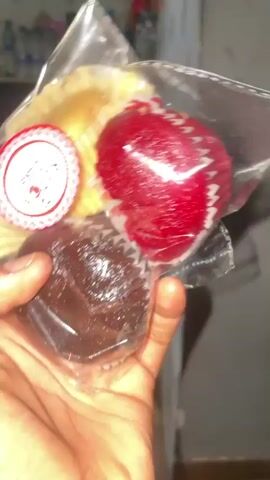 A hand holds a clear plastic bag containing three cupcakes. The cupcakes are vanilla, red velvet, and chocolate. A sticker on the bag reads “ROVOX Cake”. The background is blurry and shows various bottles and items on a surface.