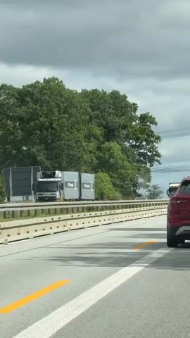 The video shows a view from a vehicle traveling on a highway. The road is paved with asphalt and marked with white lines. A yellow arrow is painted on the road in the foreground. To the left of the road, there is a concrete barrier, followed by a grassy verge and then a line of trees. On the highway, several trucks are visible. One truck, a white semi-trailer with a gray refrigerated trailer, has yellow logos on the trailer. Another truck is white with a green trailer. In the distance, more trucks and cars can be seen. The sky is cloudy.