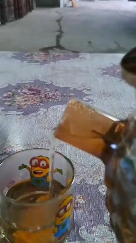 A close-up shot shows liquid being poured from a decorative brown teapot with a crystal lid into a clear glass mug. The mug features cartoon minions printed on it and already contains a yellowish liquid. The teapot is held at an angle, with its spout positioned above the mug, and a stream of clear liquid is flowing into the mug. The background is blurred but appears to be an outdoor or semi-outdoor area with a concrete floor and a wall. The table surface beneath the mug and teapot is covered with a patterned tablecloth. The lighting is natural, suggesting it is daytime.