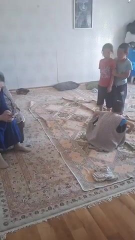 The video shows a living room scene with three children. Two children, a boy and a girl, are standing and facing towards the left of the frame, with the boy pointing. A third child is sitting on the floor facing away from the camera. The room has a patterned carpet covering most of the floor, and a blue sofa with pillows is visible in the background. There's a framed picture on the wall and a chandelier-like light fixture hanging from the ceiling. The lighting suggests it is daytime. The children appear to be playing or interacting, with one child on the floor seemingly engaged in their own activity. No visible text is present. The location context is Zhanaozen, Kazakhstan.