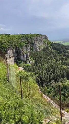 The video pans across a vista from the top of a high cliff, revealing a landscape of lush green forests, fields, and a distant town under a partly cloudy sky. The foreground features a rusty chain-link fence, bordering the cliff edge, partially obscured by vegetation. The cliff itself is a towering gray-white rock formation. In the middle ground, dense green foliage blankets the terrain, punctuated by a red-roofed building nestled amongst the trees. Further off, a town spreads out across the landscape, surrounded by fields of varying colors, including a bright yellow section. The sky above is overcast, with patches of blue peeking through. The scene has an airy, outdoor feel.