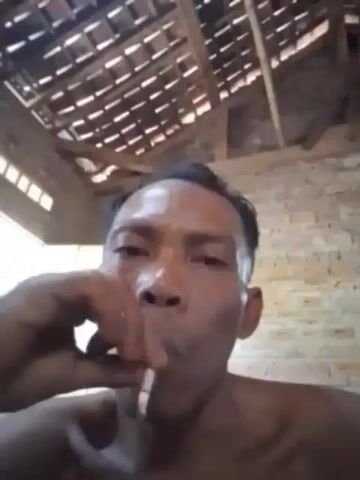 A close-up video shows a man exhaling smoke. The man is shirtless and his face is the primary focus. He appears to be indoors, with a brick wall visible in the background and a tiled roof overhead. The lighting is dim.