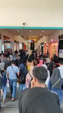 The video shows a crowded indoor walkway, likely a shopping mall. Many people, dressed casually and many wearing headscarves, are walking in the same direction, moving away from the camera. The walkway is lined with storefronts on both sides, some of which are brightly lit. There are escalators visible in the distance at the end of the walkway. A security camera is mounted high on the left wall. The lighting appears to be artificial and bright, indicating an indoor environment during the day. The overall impression is one of a busy public space.