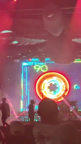 The video shows a concert or live performance. A lone male figure is visible on a stage, holding a microphone and appearing to sing or speak. Behind him, a large screen displays vibrant, abstract graphics, including a bright red circular motif that resembles a cassette tape reel with green and yellow accents. The screen also features text that reads "MINIMAL IEC TYPE 90" in neon blue and green. The stage is lit with various colored lights, including red and pink hues, and there is a haze or smoke effect creating an atmospheric ambiance. The perspective is from within the audience, with the backs of audience members' heads visible in the foreground, suggesting a crowded venue. The overall impression is a dynamic and visually engaging performance.
