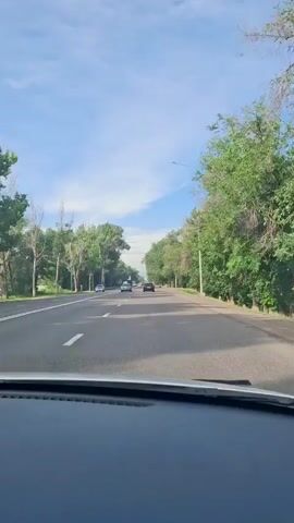 The video shows a car driving on a road lined with trees on both sides. The sky is blue with some clouds. There are other cars and a truck visible on the road ahead. The road has two lanes in each direction, separated by a double yellow line. The dashboard of the car filming the video is visible at the bottom of the frame.